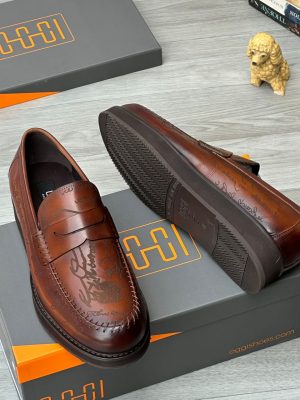 Oggi Shoes men’s brown leather penny loafers with hand-etched script design, burnished gradient finish, and rugged lug sole, displayed beside grey-and-orange branded box, poodle figurine, and stacked books on light wood surface.