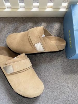 Men's luxury beige suede slip-on mules with velcro strap closure and cushioned sole on grey carpet next to blue shoebox.