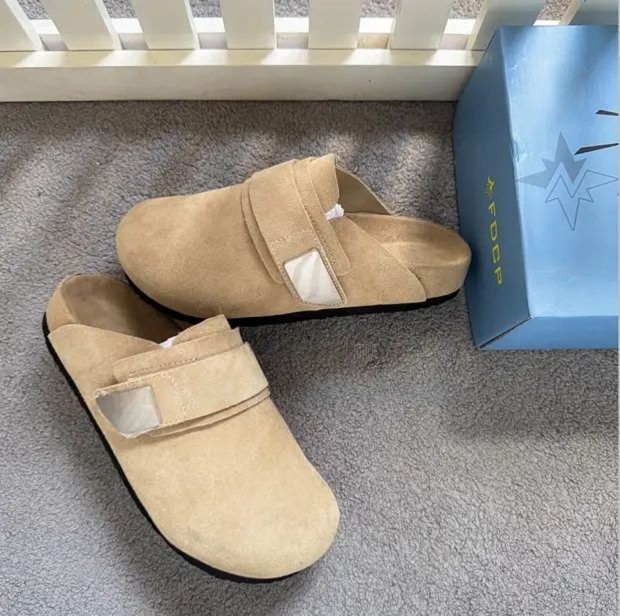 Premium beige suede slip-on mules Men's luxury beige suede slip-on mules with velcro strap closure and cushioned sole on grey carpet next to blue shoebox.