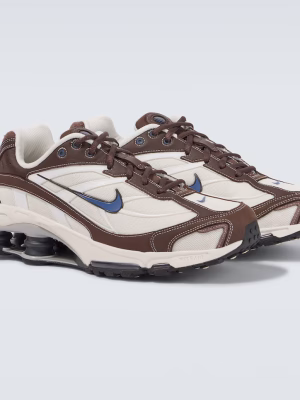 Nike Shox R4 sneakers in white, brown, and blue colorway with black Shox columns displayed on a chair against a wooden background.