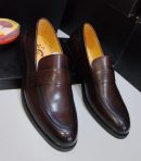 Dark brown leather penny loafers with perforated vamp detailing and tan leather lining displayed with black shoe boxes and shoe polish.