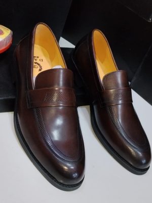 Dark brown leather penny loafers with perforated vamp detailing and tan leather lining displayed with black shoe boxes and shoe polish.