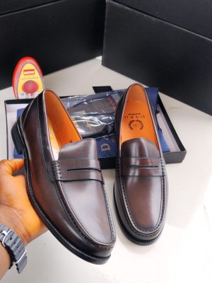 Dark brown leather penny loafers held in hand with tan lining, displayed with black shoe box and shoe polish.