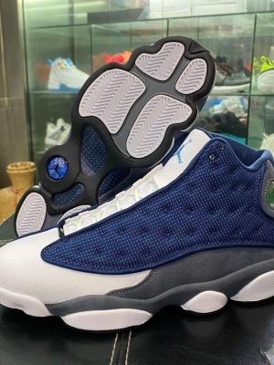 White, black, and grey Air Jordan 13 high-top sneakers with holographic eye detail and paw print outsole displayed on black box.