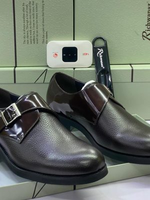 Richwanaz men’s dark brown pebbled leather monk strap shoes with patent strap and silver buckle, displayed beside branded light green box and shoe horn on white surface.