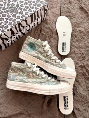 Converse x Undefeated Chuck 70 high-top sneakers in black canvas with off-white rubber sole and heel branding, held in hand with patterned shoebox and insoles visible on textured background.