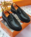 Hermès men’s black Epsom leather penny loafers with textured grain and thick rubber sole, displayed atop orange Hermès box with branded tissue paper and gold bull statue in background.