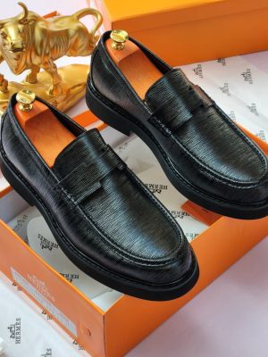 Hermès men’s black Epsom leather penny loafers with textured grain and thick rubber sole, displayed atop orange Hermès box with branded tissue paper and gold bull statue in background.