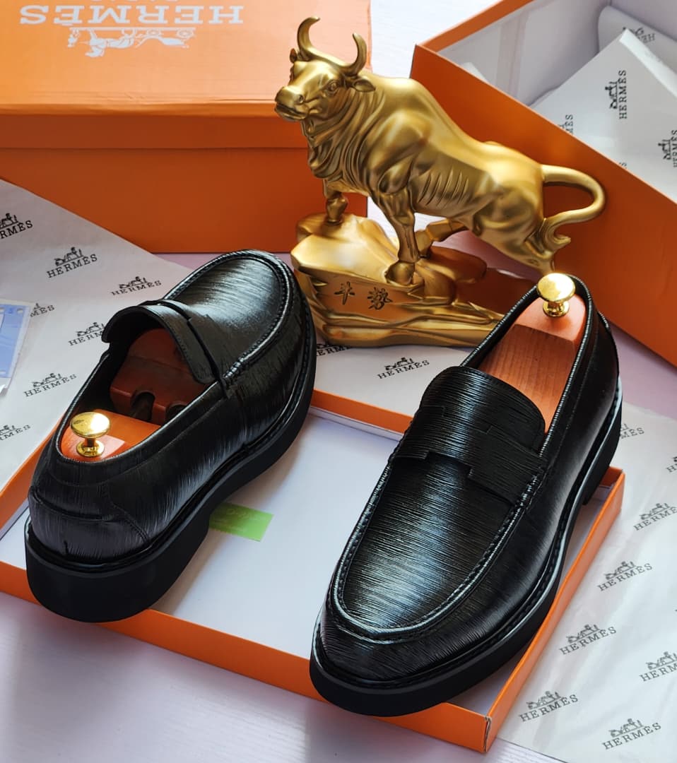 Premium Hermès black Epsom leather loafers Hermès men’s black Epsom leather penny loafers with textured grain and thick rubber sole, displayed atop orange Hermès box with branded tissue paper and gold bull statue in background.