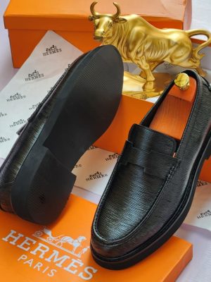 Hermès men’s black Epsom leather penny loafers with textured grain and thick rubber sole, displayed atop orange Hermès box with branded tissue paper and gold bull statue in background.