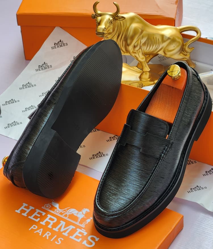 Premium Hermès black Epsom leather loafers Hermès men’s black Epsom leather penny loafers with textured grain and thick rubber sole, displayed atop orange Hermès box with branded tissue paper and gold bull statue in background.