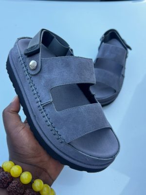 UGG men's grey suede fisherman sandals with double velcro straps and contrast stitching, held in hand against white car background.
