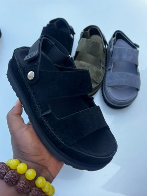 UGG men's grey suede fisherman sandals with double velcro straps and contrast stitching, held in hand against white car background.