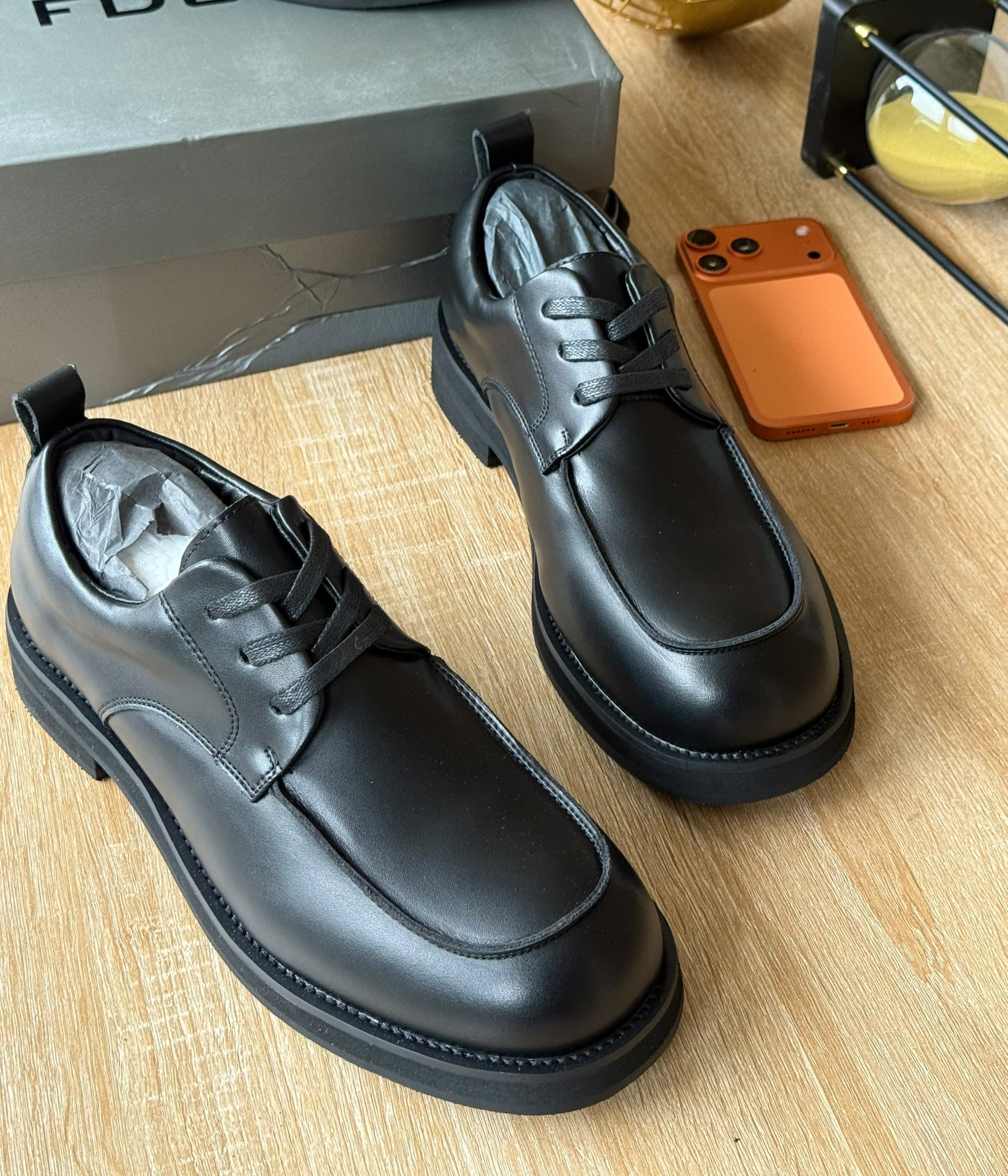 Premium FDCP black leather derby shoes FDCP men's black leather derby shoes with tonal laces and thick black rubber sole, displayed on wooden surface with grey branded shoebox and orange phone.
