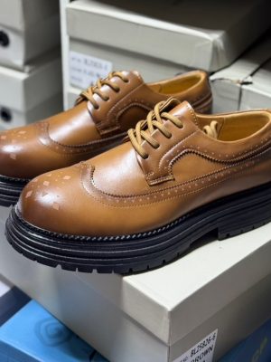Men's luxury cognac brown leather brogue oxford shoes with geometric toe cap detailing and thick black ridged sole sitting on white shoeboxes.