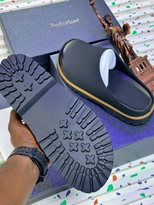 Timberland men's black leather clog mules with tan stitching and rubber lug sole, held in hand with branded box and Statue of Liberty figurine on terrazzo background.
