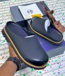 Timberland men's black leather clog mules with tan stitching and rubber lug sole, held in hand with branded box and Statue of Liberty figurine on terrazzo background.