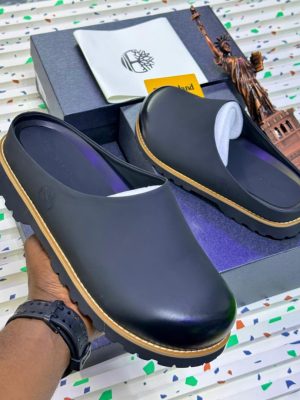 Timberland men's black leather clog mules with tan stitching and rubber lug sole, held in hand with branded box and Statue of Liberty figurine on terrazzo background.