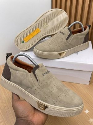 Timberland men's beige suede slip-on sneakers with perforated upper, cream rubber sole, and gold logo plaque, displayed with white shoebox on wooden surface.