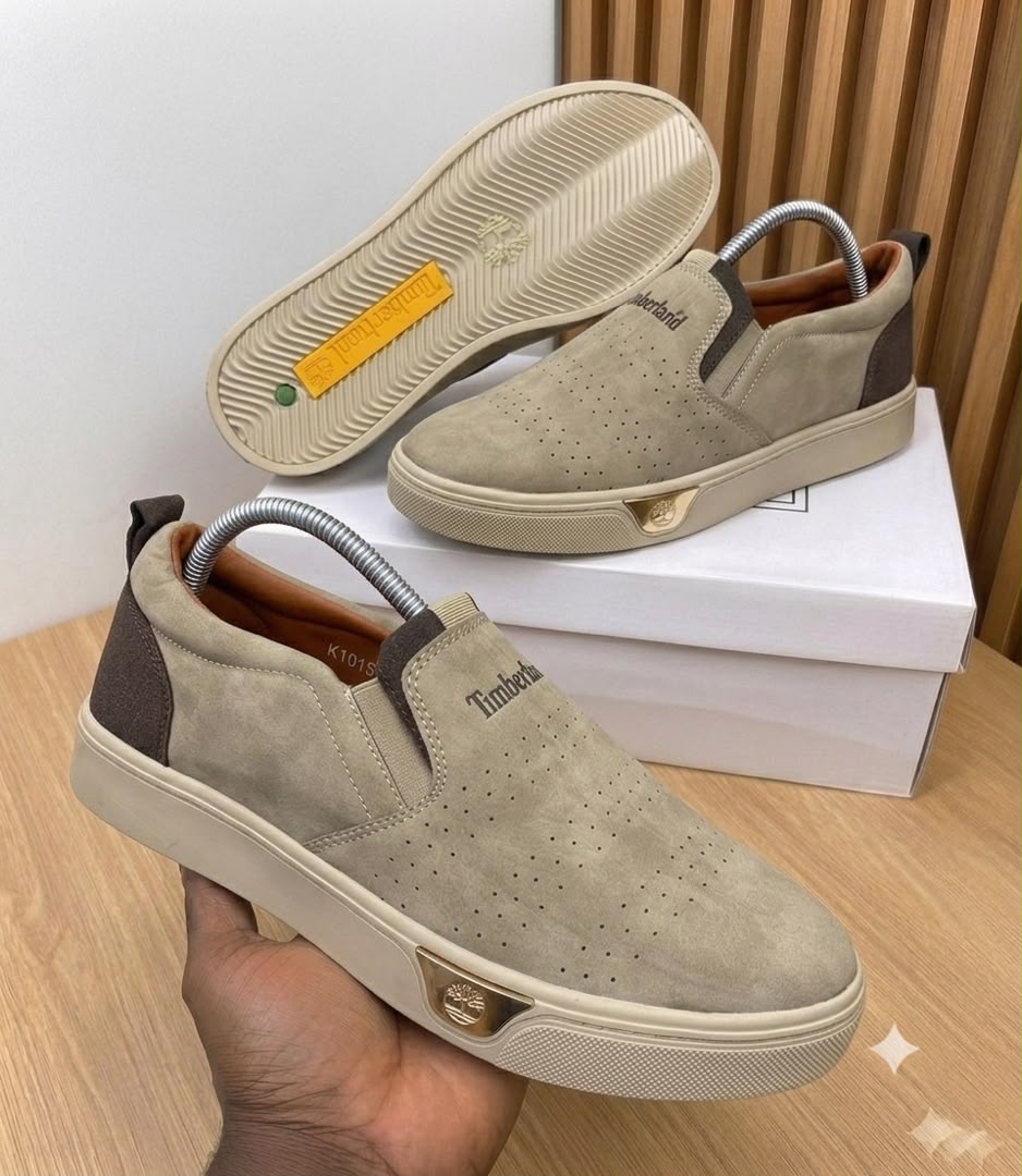 Timberland beige suede slip-on sneakers Timberland men's beige suede slip-on sneakers with perforated upper, cream rubber sole, and gold logo plaque, displayed with white shoebox on wooden surface.