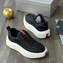 Prada men's black mesh platform sneakers with rubberized overlays, white chunky sole, and red heel tab, displayed on a grey wood floor with a silver shoebox and gold apple decor.