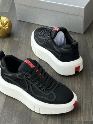 Prada men's black mesh platform sneakers with rubberized overlays, white chunky sole, and red heel tab, displayed on a grey wood floor with a silver shoebox and gold apple decor.