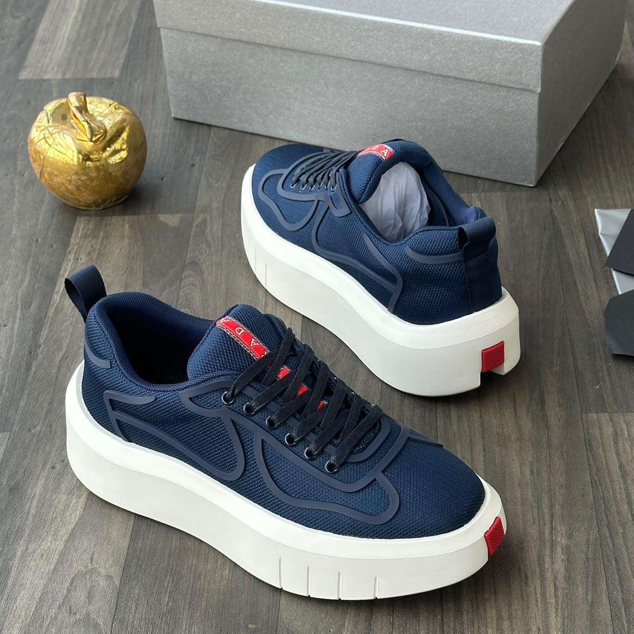 Prada black mesh platform sneakers Prada men's black mesh platform sneakers with rubberized overlays, white chunky sole, and red heel tab, displayed on a grey wood floor with a silver shoebox and gold apple decor.