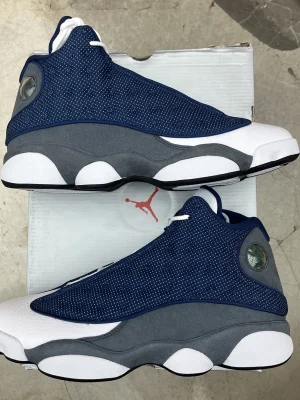 White, black, and grey Air Jordan 13 high-top sneakers with holographic eye detail and paw print outsole displayed on black box.