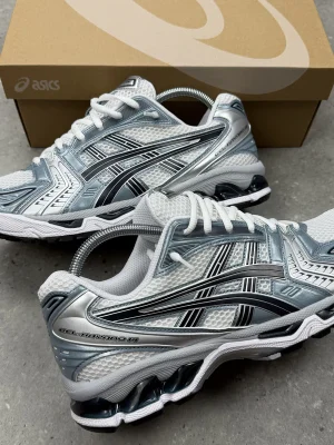 ASICS GEL-Kayano 14 men's sneakers in silver, white, and blue with visible GEL cushioning units, displayed on grey concrete surface with open cardboard shoebox.