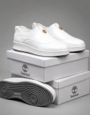 Timberland men's white leather slip-on sneakers with gold medallion and thick white rubber sole with grey tread, displayed on stacked white shoeboxes.