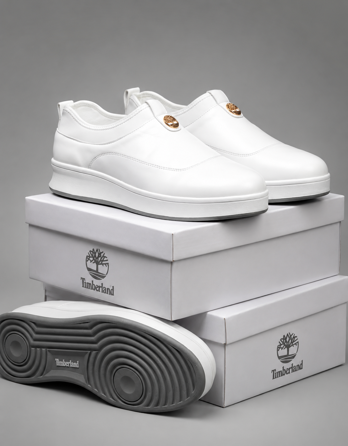 Timberland white leather slip-on sneakers Timberland men's white leather slip-on sneakers with gold medallion and thick white rubber sole with grey tread, displayed on stacked white shoeboxes.