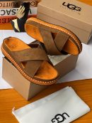Black UGG suede cross-strap slides with white contrast stitching and chunky sole displayed with UGG box and dust bag.