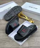 Black Miu Miu leather slides with red and black embroidered logo and chunky treaded sole displayed in white box with gold decor.