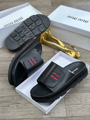 Black Miu Miu leather slides with red and black embroidered logo and chunky treaded sole displayed in white box with gold decor.