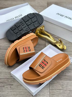 Black Miu Miu leather slides with red and black embroidered logo and chunky treaded sole displayed in white box with gold decor.