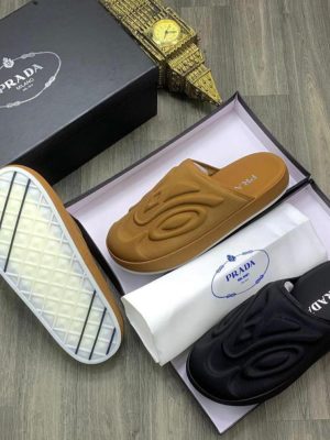 Black and camel Prada slides with large embossed logo displayed in box with dust bag and gold clock decor.