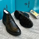 Steve Riots men’s black leather cap-toe brogue boots with glossy finish, perforated detailing, and rugged lug sole, displayed beside turquoise branded box and metallic VIP tag on textured grey surface.