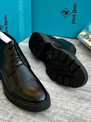 Steve Riots men’s black leather cap-toe brogue boots with glossy finish, perforated detailing, and rugged lug sole, displayed beside turquoise branded box and metallic VIP tag on textured grey surface.