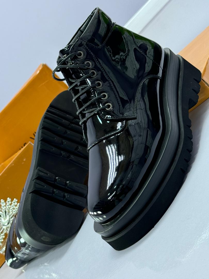 Premium Louis Vuitton combat boots Black patent leather combat boots with chunky platform sole and lace-up front, displayed beside orange Louis Vuitton box and decorative perfume bottle.