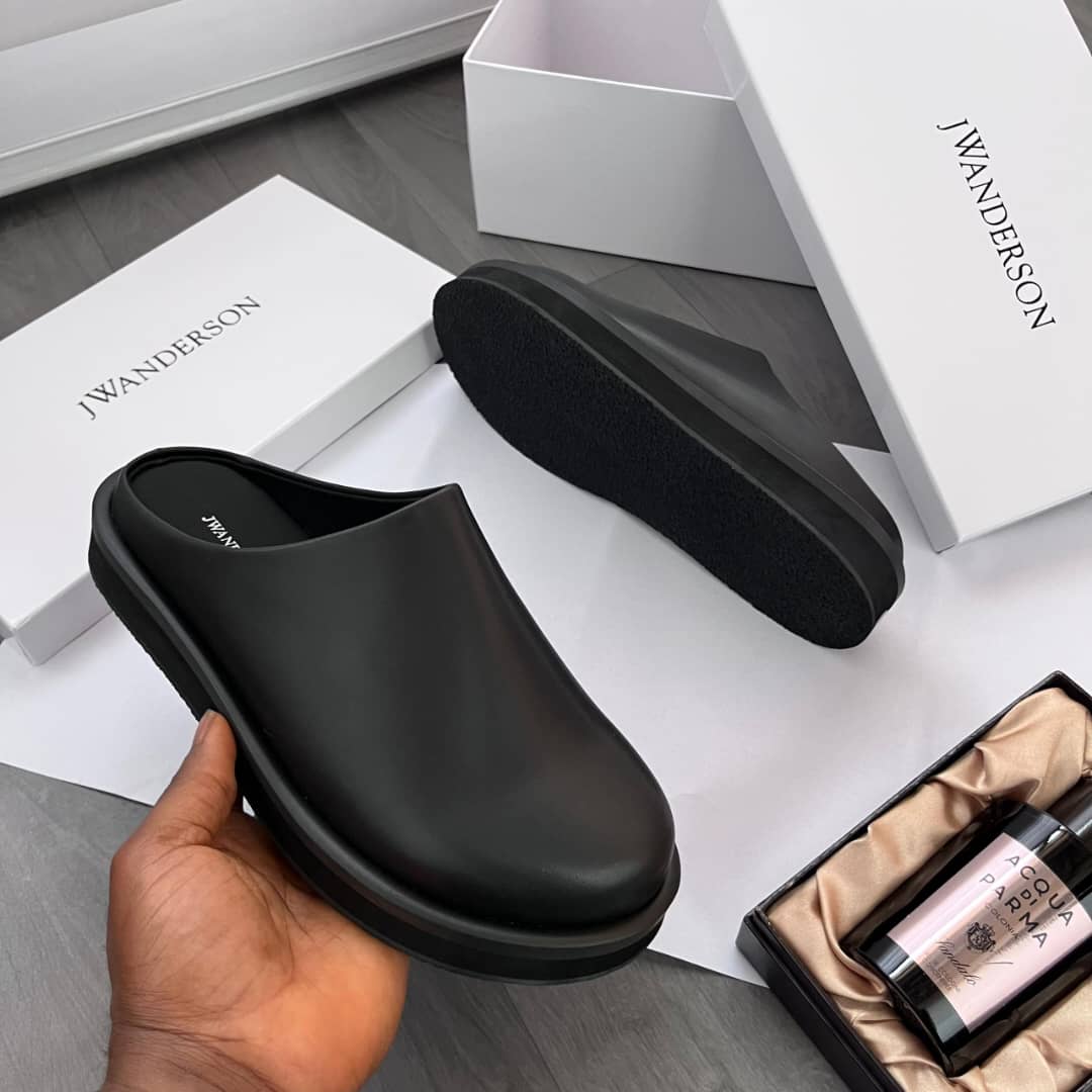 JW Anderson leather slip-on mules JW Anderson men’s brown leather slip-on mules with sculptural silhouette, two-tone platform sole, and branded insole, held by hand beside white branded box and Acqua di Parma gift set on grey wood floor.