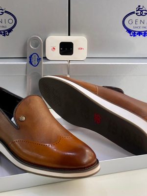 Genio men’s brown leather brogue loafers with burnished finish, wingtip perforations, white contrast sole, and red “G” logo, displayed beside branded silver box and electronic device on white surface.