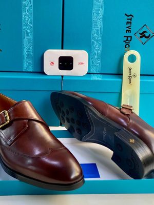 Steve Riots men’s brown leather monk strap shoes with burnished gradient finish, gold-tone buckle, and rugged lug sole, displayed beside turquoise branded box and electronic device on white surface.