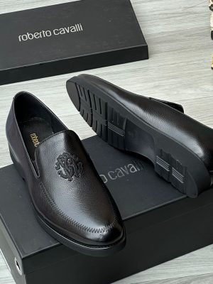 Roberto Cavalli men’s brown textured leather loafers with embossed RC monogram and rugged lug sole, displayed beside branded black box, poodle figurine, and stacked books on light wood surface.