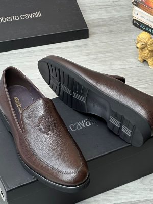 Roberto Cavalli men’s brown textured leather loafers with embossed RC monogram and rugged lug sole, displayed beside branded black box, poodle figurine, and stacked books on light wood surface.