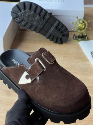 Alexander McQueen men’s brown suede slip-on sandals with silver triangular buckle and thick black lug sole, displayed with branded white box and dust bag on wooden surface.