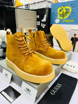 Balenciaga men's mustard yellow suede combat boots with tonal laces, side zipper, and thick crepe sole, displayed on grey shoebox with branded cards and accessories.
