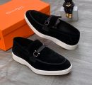 Santoni men's black suede loafer sneakers with tonal leather strap, silver hardware, and white rubber sole, displayed with orange branded shoebox on grey wood floor.