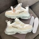 Men's luxury white Balenciaga Triple S sneakers with beige accents and distinctive clear transparent soles resting on a grey sofa.
