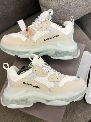 Men's luxury white Balenciaga Triple S sneakers with beige accents and distinctive clear transparent soles resting on a grey sofa.