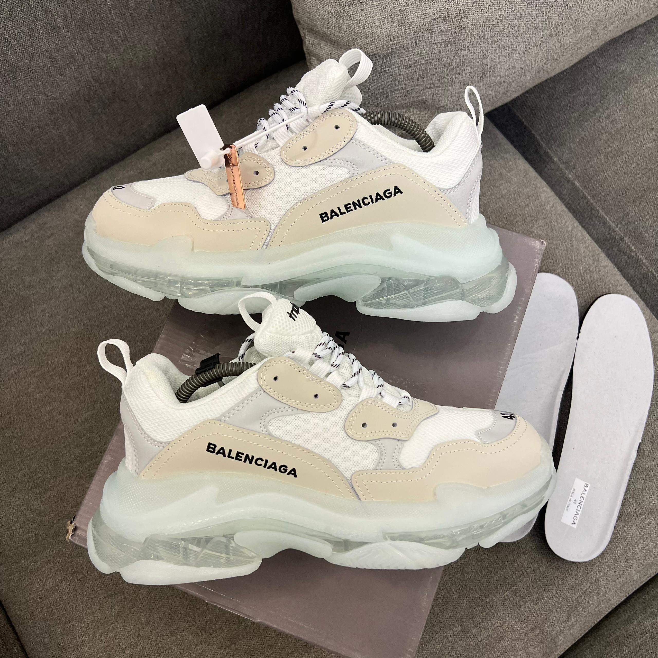 Iconic Balenciaga Triple S sneakers Men's luxury white Balenciaga Triple S sneakers with beige accents and distinctive clear transparent soles resting on a grey sofa.